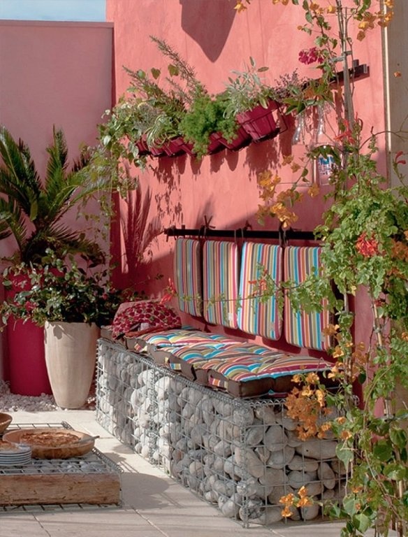 a bright outdoor Mediterranean space with a coral wall, a built in bench with pebbles inside, bright striped upholstery and lots of greenery around is very atmospheric