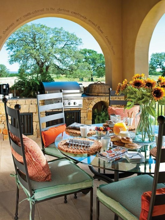 an outdoor Mediterranean dining space with an outdoor kitchen clad with stone, with a glass table and green chairs with red pillows