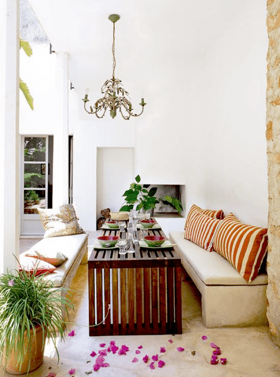 an indoor-outdoor dining space with a Mediterranean feel, with a fireplace, neutral upholstered benches, a stained wooden table and potted greenery