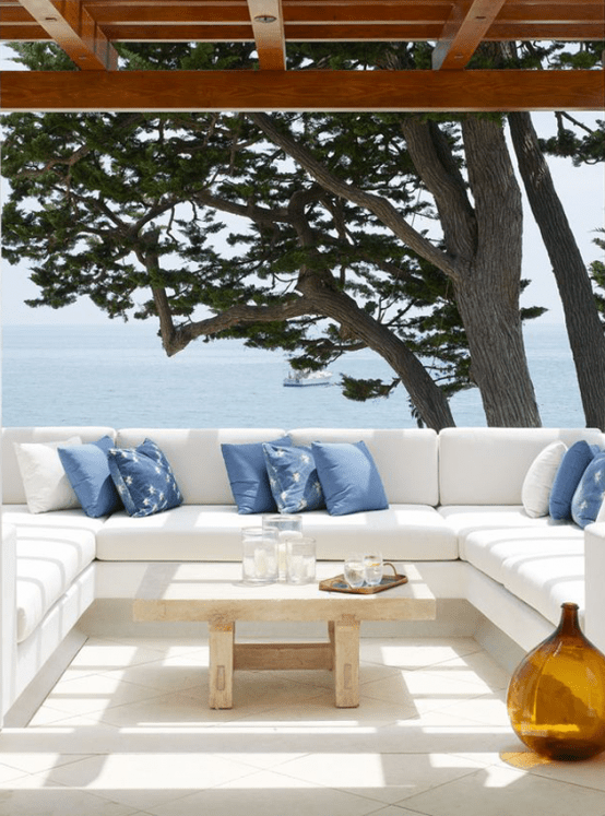 a lovely Mediterranean outdoor space with a built-in sofa with white upholstery, blue pillows, a low table and an amber bottle plus a gorgeous sea view