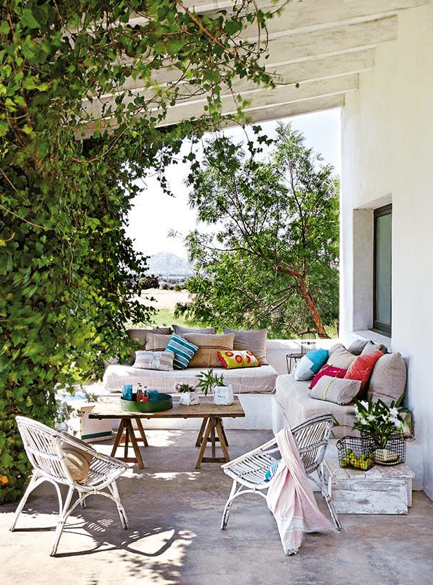 a lively Mediterranean outdoor space with a white built in sofa with neutral upholstery and bright pillows, a wooden trestle table and wicker chairs, greenery weaving around the pillars