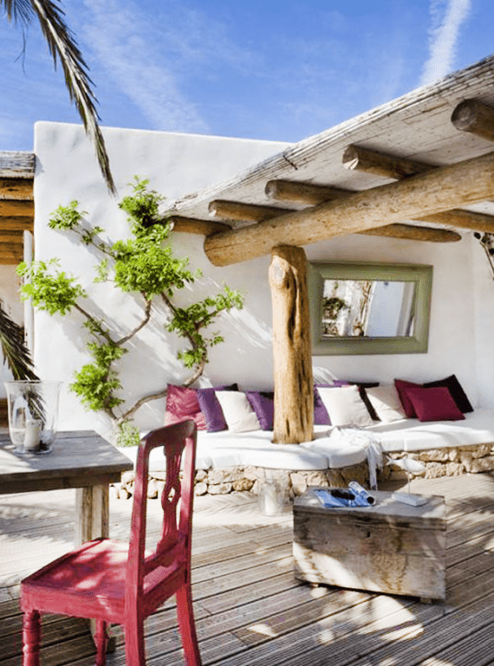 a pretty Mediterranean outdoor space with a roof over a built-in sofa with white upholstery, a wooden side table, a wooden table and a pink chair, some greenery