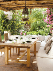 a white Mediterranean outdoor space with a built-in white sofa, a wooden table, woven chairs, a Moroccan pendant lamp and bright blooms around