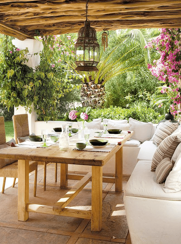 a white Mediterranean outdoor space with a built in white sofa, a wooden table, woven chairs, a Moroccan pendant lamp and bright blooms around