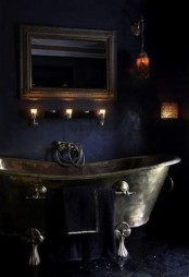 a refined and dark Gothic bathroom with navy walls, a vintage metal bathtub, various vintage lamps, a mirror in a vintage frame and a dark floor
