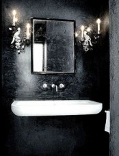 a beautiful Gothic bathroom with textural black walls, a floating white sink, candlelabra-styled lamps and a mirror in a modern black frame
