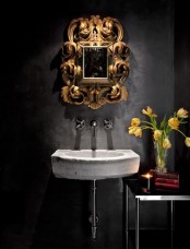 a dramatic bathroom with graphite grey walls, a concrete floating sink, a mirror with an oversized gold frame, a small coffee table