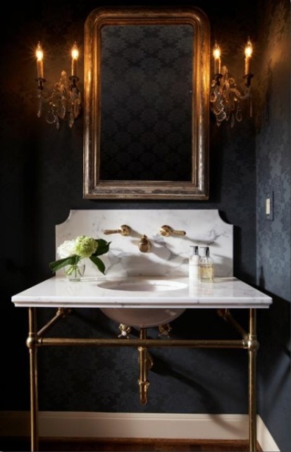 a vintage Gothic bathroom with black printed wallpaper, a free-standing sink of white marble, candelabra-style lamps and a mirror in a gilded frame