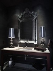 a Gothic bathroom with black walls, a mirror with an ornated frame, chic table lamps and a built-in sink