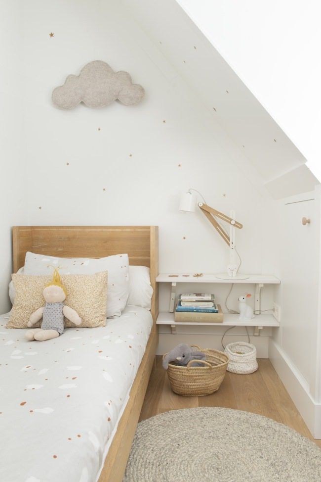 a delicate and subtle Scandinavian kid's room with a light stained bed and neutral bedding, an open shelf nightstand, a cloud decoration and a white sconce and pastel bedding