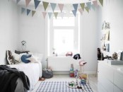 a small Scandi kid’s room with sleek white furniture, pastel-colored buntings, ledges with books and artwork, a printed rug and lots of toys