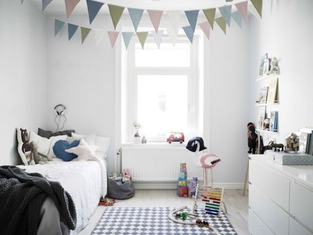 a small Scandi kid's room with sleek white furniture, pastel colored buntings, ledges with books and artwork, a printed rug and lots of toys