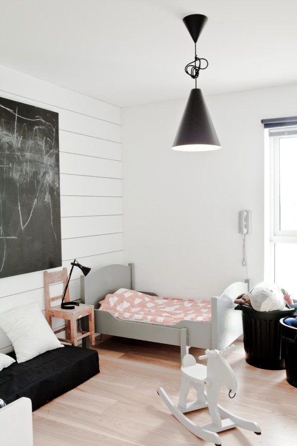 a welcoming Scandinavian kid's room with a white shiplap wall, a grey bed with pastel bedding, black baskets for storage, a chalkboard and a low sofa with pillows
