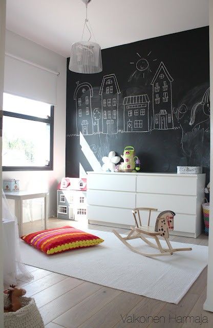 a Nordic kid's room with a black chalkboard wall, a white sleek dresser, white furniture and lots of different toys, a creamy rug