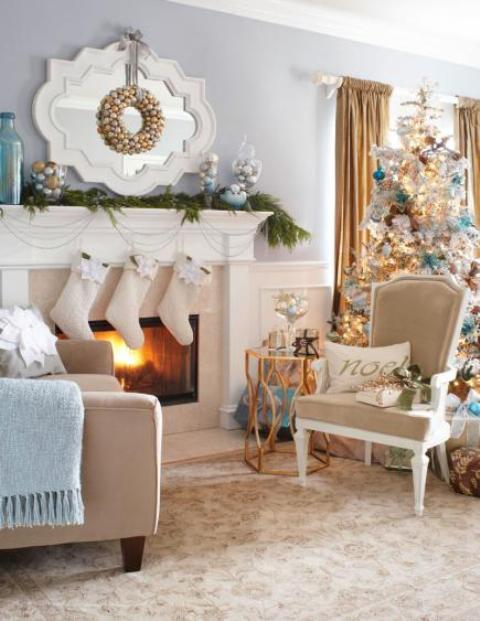 white stockings, beads, fir garlands on the mantel, a gold ornament wreath, a flocked Christmas tree with blue and gold ornaments for refined holiday decor