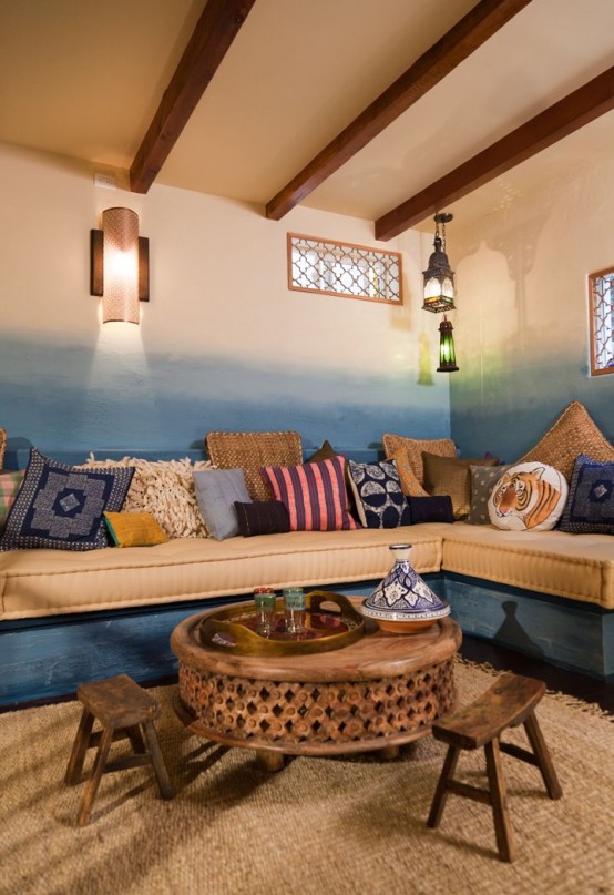a Moroccan living room with ombre blue walls, a built-in sofa, colorful and printed pillows, a carved wood table and stools