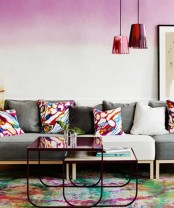 a bold interior with a pink ombre accent wall, a grey sofa with bright pillows, a couple of burgundy coffee tables and red drip pendant lamps