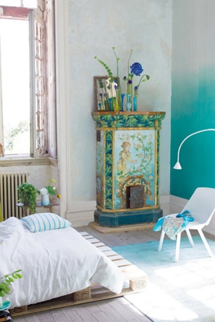 a bold bedroom with an ombre green wall, a green floral fireplace, a pallet bed with neutral bedding, potted blooms and greenery