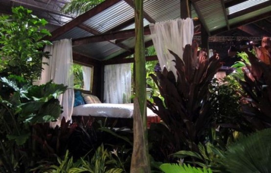 a garden bedroom with a roof featuring a canopy bed, greenery and colorful foliage and neutral curtains