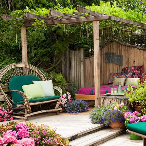 a colorful outdoor boho space with a bed under a roof with greenery, with colorful bedding, a rattan chair with pillows and lots of blooms around
