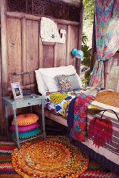 a colorful boho outdoor bedroom with a wooden wall, a bed with colorful bedding, a nightstand, colorful pillows and curtains and a rug on the floor