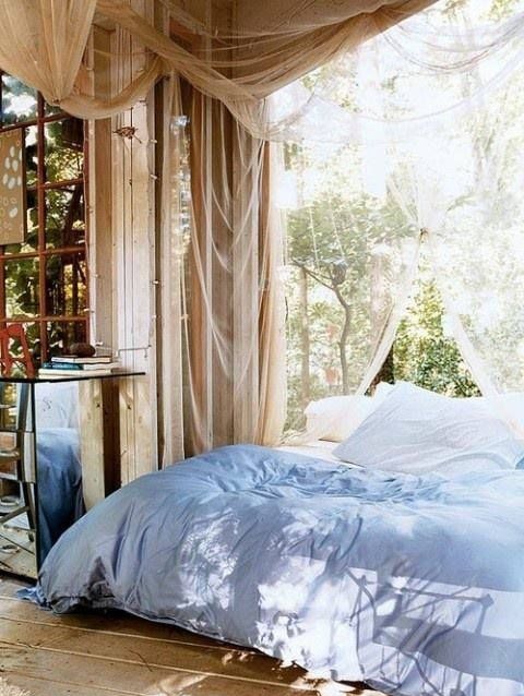 a neutral outdoor indoor bedroom with mosquito nets as curtains, a bed with pastel blue bedding and lots of greenery around