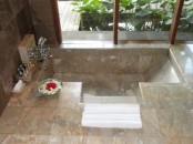 a natural-looking bathroom with neutral stone tiles and a sunken bathtub with steps and a glazed wall with a view