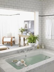 a sunken bathtub clad with neutral stone and with a window to the bedroom plus some candles and a potted plant