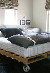 a modern industrial bedroom with a pallet bed on casters, white and grey bedding, a whitewashed dresser, printed curtains
