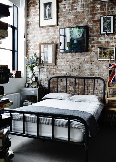 an industrial bedroom with brick walls, a black metal bed, mismatching nightstands, a free form gallery wall and bookshelves