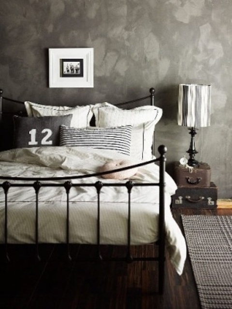 a moody bedroom with a black metal bed with neutral bedding, stacked suitcases and a table lamp is a relaxing space