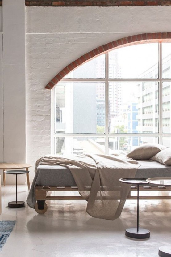 a neutral bedroom with an arched window, pallet bed with grey bedding, metal and wooden coffee and side tables