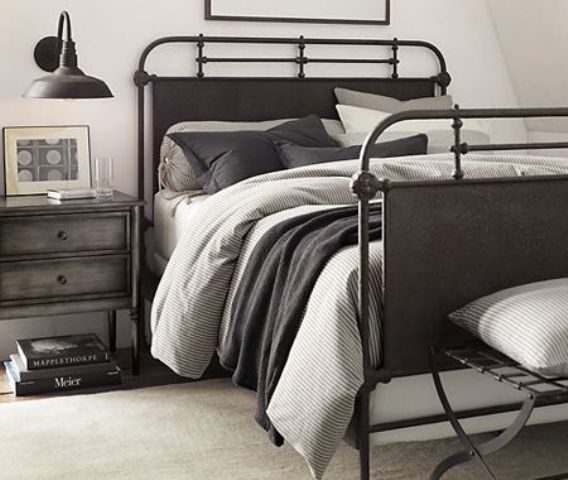 an industrial bedroom with a blackened metal bed and a metal bench, grey and black bedding, a grey nightstand and a black sconce