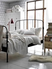 an industrial bedorom clad with white square tiles, with metal cabinets, a blackened metal bed with white bedding, a red table lamp and stacks of books