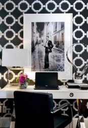 an elegant black and white home office with printed wallpaper, a white desk, black chair, chic artworks and a white table lamp