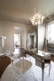 a refined neutral home office with printed wallpaper, a tall mirror in a frame, a mirrored desk and an acrylic chair plus a statement chandelier