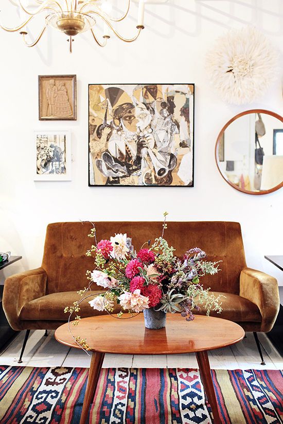 a boho mid-century modern space with a mustard mid-century modern loveseat, a bright printed rug, a stained table and a gallery wall with a round mirror plus a chic chandelier