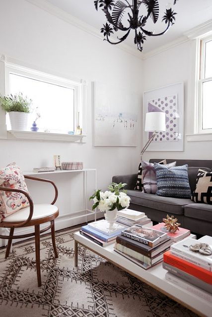 a small bright living room with eclectic furniture, a printed rug, a black chandelier, a coffee table with lots of books and magazines