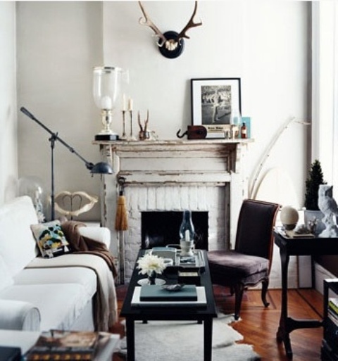 a small eclectic living room with a non-workign fireplace, vintage furniture, antlers, artworks and lamps and candles