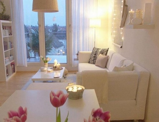 a small white living room with comfy furniture, lamps and chandeliers, string lights and a glazed wall