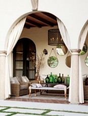a neutral rustic terrace with a wicker sofa, a daybed with pillows, a coffee table with an arrangement of bottles and neutral curtains