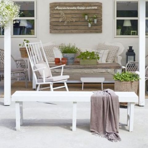 a rustic neutral terrace with a grey sofa, a whiet rocker and a footrest, a white bench and a taupe blanket, potted greenery here and there