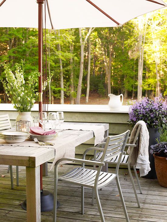a neutral rustic terrace with a stained wooden table, metal chairs, an umbrella and potted blooms around