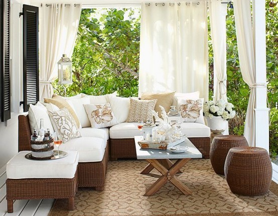 a small and cozy neutral terrace with brown wicker furniture with white upholstery, a coffee table and neutral curtains around