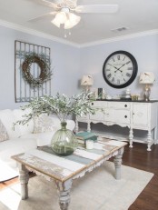 a shabby chic living room with pastel blue walls, refined neutral furniture, a wreath and a clock, a low table and neutral linens
