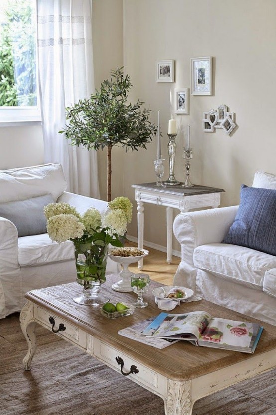 a refined shabby chic living room in neutrals, with white furniture, a low table, blue pillows and potted greenery