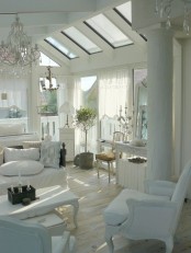 a white shabby chic living room with skylights, elegant white furniture, a crystal chandelier, a potted plant and greenery