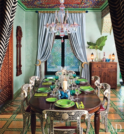 a bright Moroccan-inspired dining room with a bright checked floor, a bold ceiling, a heavy carved table and painted chairs plus bold printed textiles