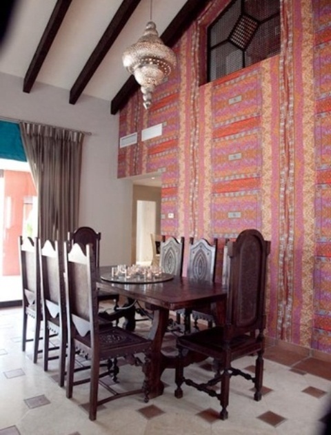 a pretty Moroccan dining room with a wall covered with bold tapestries, heavy carved wooden furniture, a metal pendant lamp and wooden beams