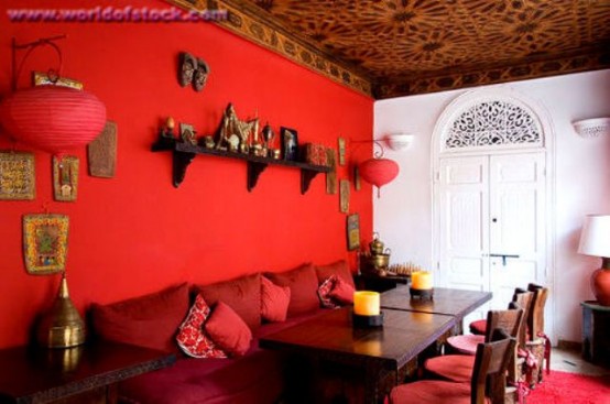 a bold red Moroccan dining room with a hot red accent wall, a shelf and red paper lamps, a dark table and an upholstered bench, a patterned ceiling and red chairs
